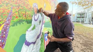 Jorge Arce takes a small paintbrush to paint details on a woman in a traditional-style white dress for la Bamba dance.